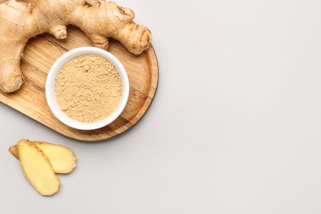 Wooden board with fresh ginger root and bowl of dried powder on gray background