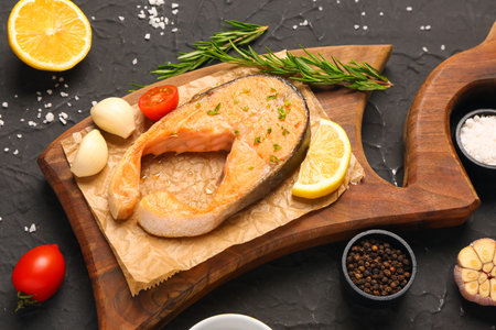 Wooden board of tasty salmon steak with tomatoes, garlic and lemon on black background