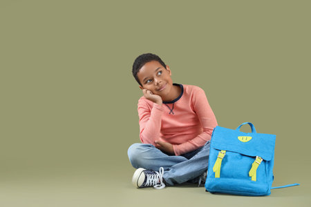 Dreaming African-American boy sitting near school backpack on green backgroundの写真素材