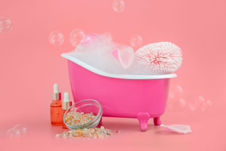 Small bathtub with foam, sponge and cosmetic products on color background