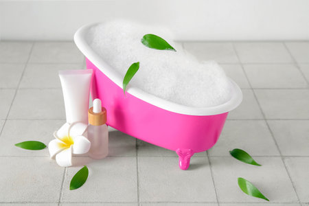 Small bathtub with foam, cosmetic products and plant leaves on light tile tableの写真素材