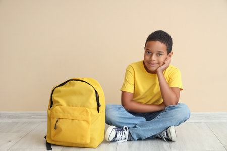 Little African-American boy sitting near school backpack on beige backgroundの写真素材