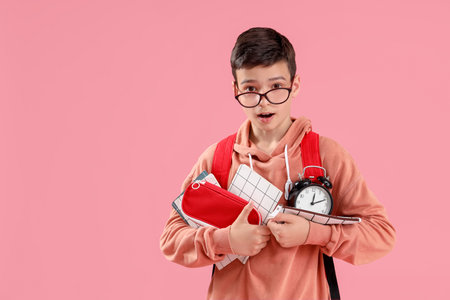 Portrait of shocked schoolboy with school supplies and alarm clock on pink backgroundの写真素材