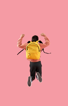 Jumping schoolboy with yellow backpack on pink background, back viewの写真素材