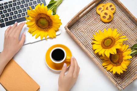 Woman with cup of coffee, modern laptop, sunflowers and cookies on light backgroundの写真素材