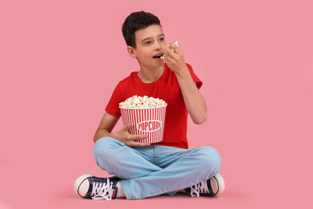 Cute little boy eating popcorn while sitting on pink backgroundの写真素材