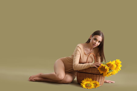 Young woman with wicker basket full of beautiful sunflowers sitting on green backgroundの写真素材