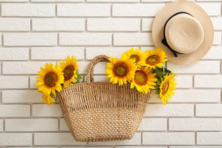 Stylish straw bag with hat and beautiful sunflowers on white brick wallの写真素材