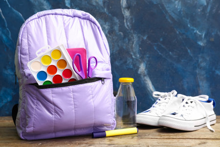 Lilac school backpack with watercolors, bottle of water and sneakers on wooden table near blue grunge wallの写真素材