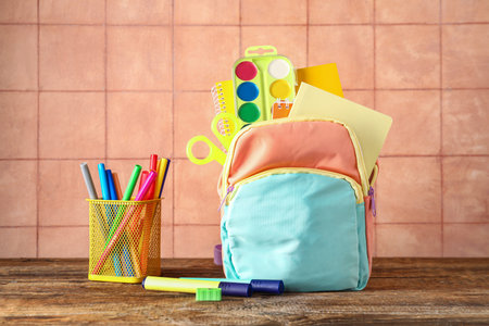 Colorful school backpack with notebooks, watercolors and cup of markers on wooden table near beige tile wallの写真素材