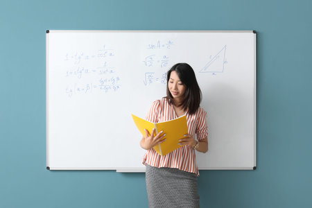 Asian Math teacher with notebook near flipboard in classroom
