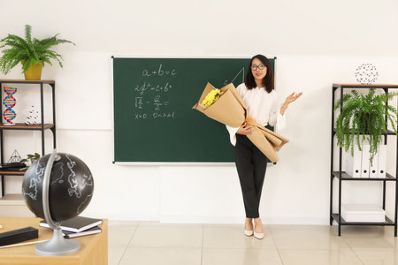 Female Asian teacher with bouquet of flowers near chalkboard in classroomの写真素材
