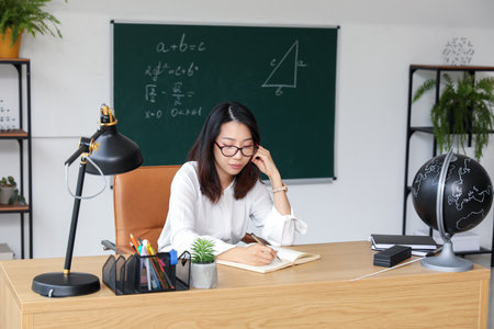 Female Asian teacher writing at table in classroomの写真素材