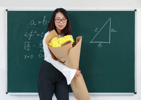 Female Asian teacher with bouquet of flowers near chalkboard in classroomの写真素材