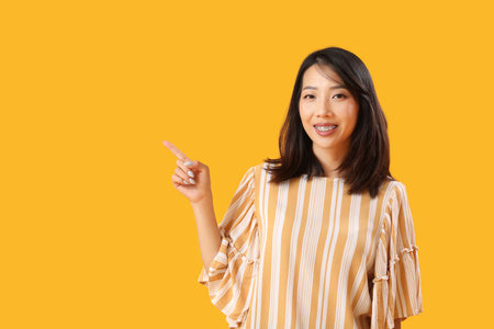 Beautiful Asian woman pointing at something on yellow background. Dialogue conceptの写真素材