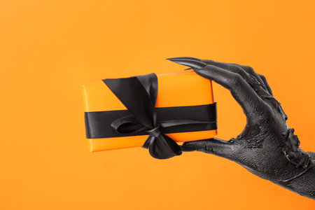 Black hand of witch with gift box on orange background, closeup. halloween celebrationの写真素材