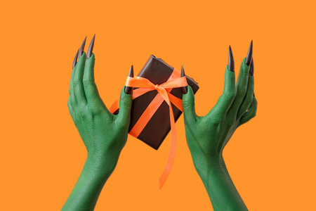 Green hands of witch with gift box on orange background. halloween celebrationの写真素材
