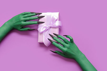 Green hands of witch with gift box on purple background. halloween celebration