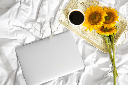 Beautiful sunflowers, cup of coffee, eyeglasses and modern laptop on light fabric backgroundの写真素材