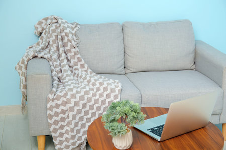 Gray sofa with cozy blanket and laptop on coffee table near blue wall