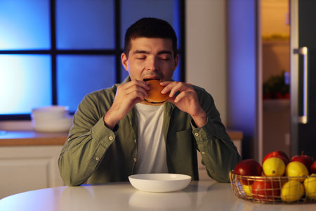 Hungry young man eating burger in kitchen at nightの写真素材