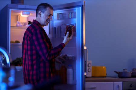 Hungry mature man with jam near open fridge in kitchen at nightの写真素材