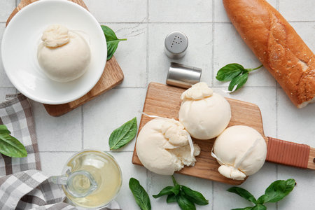 Wooden board and plate of tasty Burrata cheese with basil on white tile background