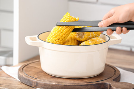 Woman taking out boiled corn cobs from cooking pot on wooden table in kitchenの写真素材