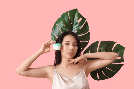 Beautiful young Asian woman with facial cream and tropical leaves on pink backgroundの写真素材
