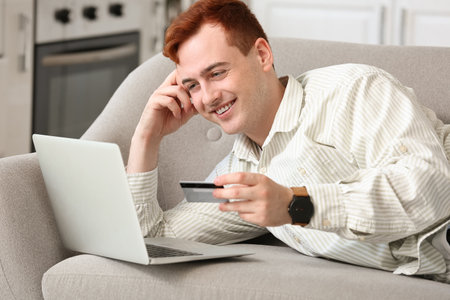 Handsome young man with credit card and modern laptop lying on sofa while shopping onlineの写真素材