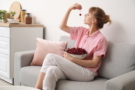 Beautiful happy young woman sitting on sofa and eating ripe cherries in kitchenの写真素材