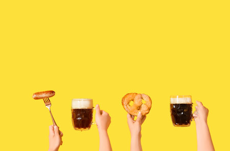 Female hands with mugs of cold beer, sausage and pretzel on yellow backgroundの写真素材