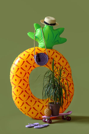 Inflatable ring in shape of pineapple with skateboard and beach accessories on green background....