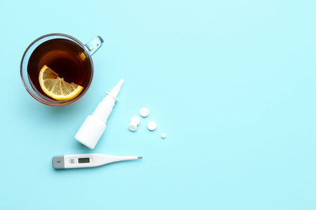 Cup of tea, pills, nasal spray and thermometer on blue background