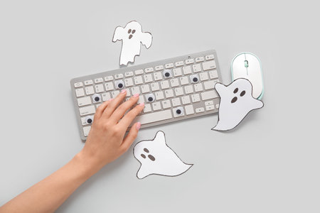 Female hand with modern computer keyboard, mouse and paper ghosts for Halloween on gray backgroundの写真素材
