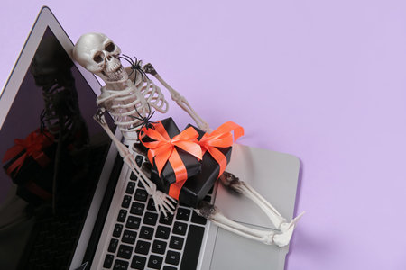 Composition with modern laptop, skeleton and gift boxes for Halloween on purple background, closeupの写真素材