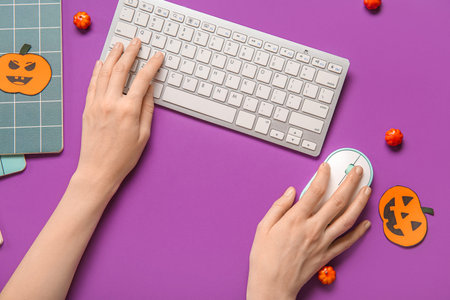 Female hands with modern computer keyboard, mouse, notebook and pumpkins for Halloween on purple...