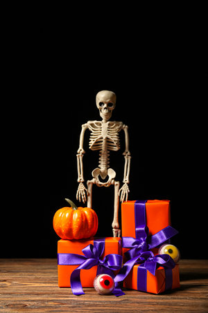 Beautiful gift boxes, skeleton, pumpkin and tasty candies for Halloween on wooden table against dark backgroundの写真素材