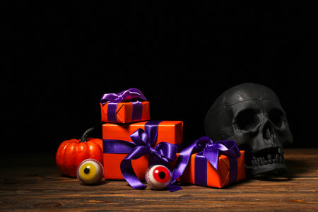 Beautiful gift boxes, skull and tasty candies for Halloween on wooden table against dark backgroundの写真素材