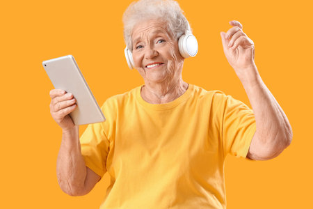 Senior woman in headphones with tablet computer listening to music on orange background