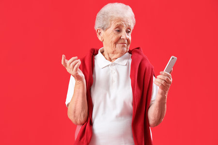 Confused senior woman with mobile phone on red backgroundの写真素材