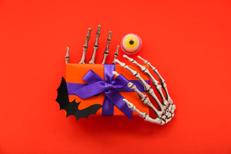 Composition with skeleton hands, gift box and tasty candy for Halloween on red backgroundの写真素材