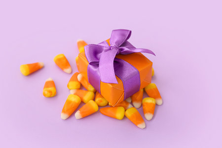 Gift box and tasty candy corns for Halloween on purple background, closeup