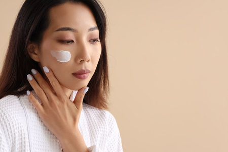 Beautiful Asian woman with cream on her face against beige backgroundの写真素材
