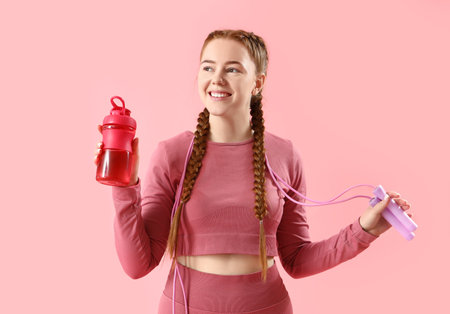 Sporty young woman with skipping rope and bottle of water on pink backgroundの写真素材