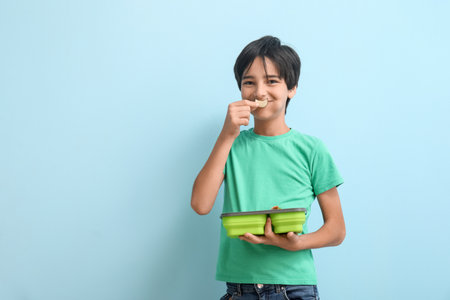 Little boy with lunchbox eating cucumber slice on blue backgroundの写真素材