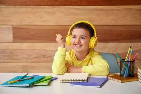 Cute little boy in headphones doing homework at table near wooden wallの写真素材