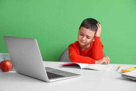 Stressed little boy doing homework at table near green wallの写真素材