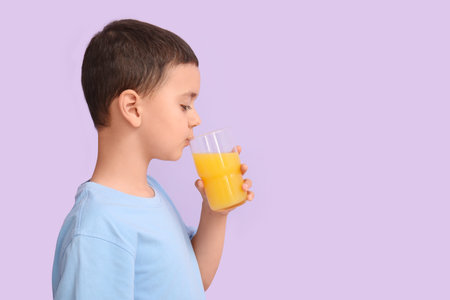 Cute little boy drinking orange juice on lilac backgroundの写真素材