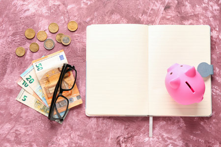 Blank notebook with piggy bank, money and eyeglasses on grunge pink backgroundの写真素材